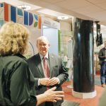 Senator Coons in a black suit with a red tie speaks to woman with her back turned to camera