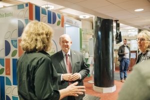 Senator Coons in a black suit with a red tie speaks to woman with her back turned to camera
