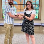 Man with brown beard wearing a white and blue stripped shirt shaking hands with woman with brown, curly hair with black glasses wearing a black and white polka dotted dress
