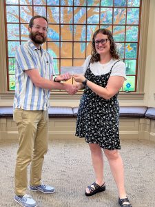 Man with brown beard wearing a white and blue stripped shirt shaking hands with woman with brown, curly hair with black glasses wearing a black and white polka dotted dress